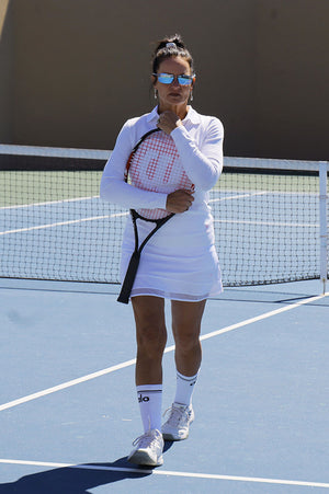 Woman playing tennis in a white skort with mesh trim and a white polo and white sweater holding a tennis racket