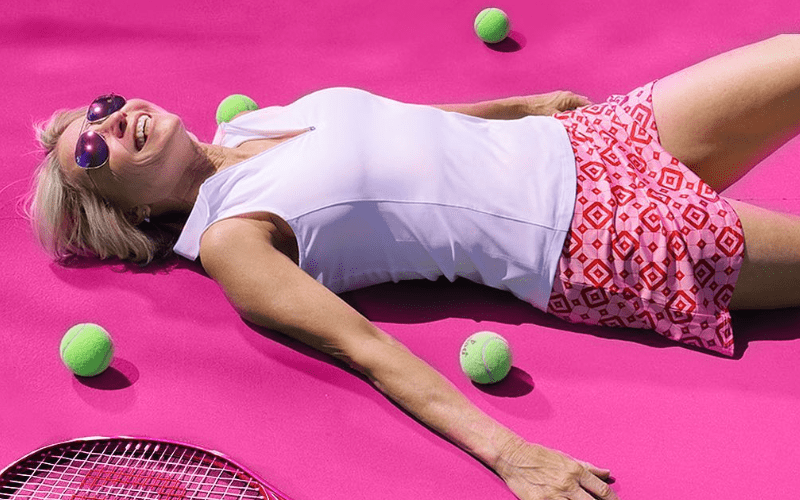 A woman in a tennis outfit lying on a pink court, surrounded by tennis balls.